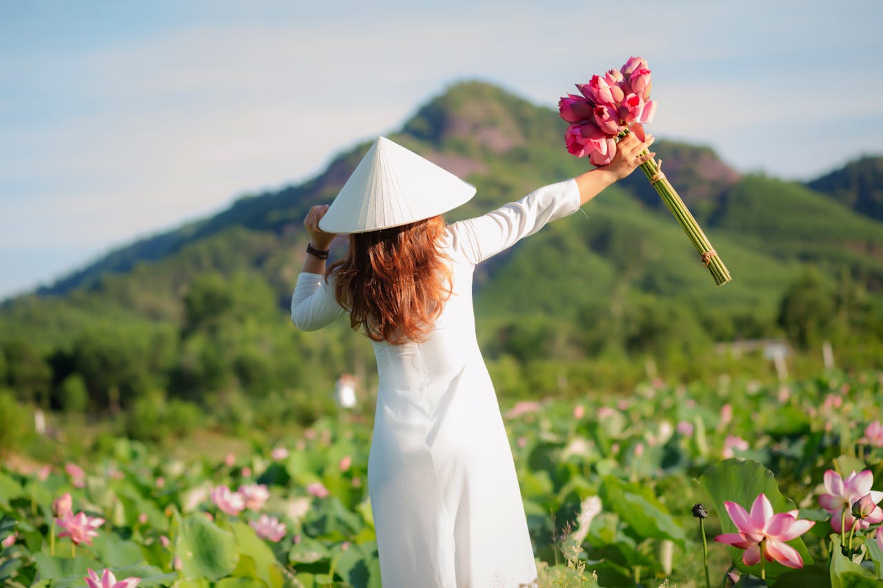 Woman in traditional ao dai holding lotus flowers in a scenic Vietnamese landscape.